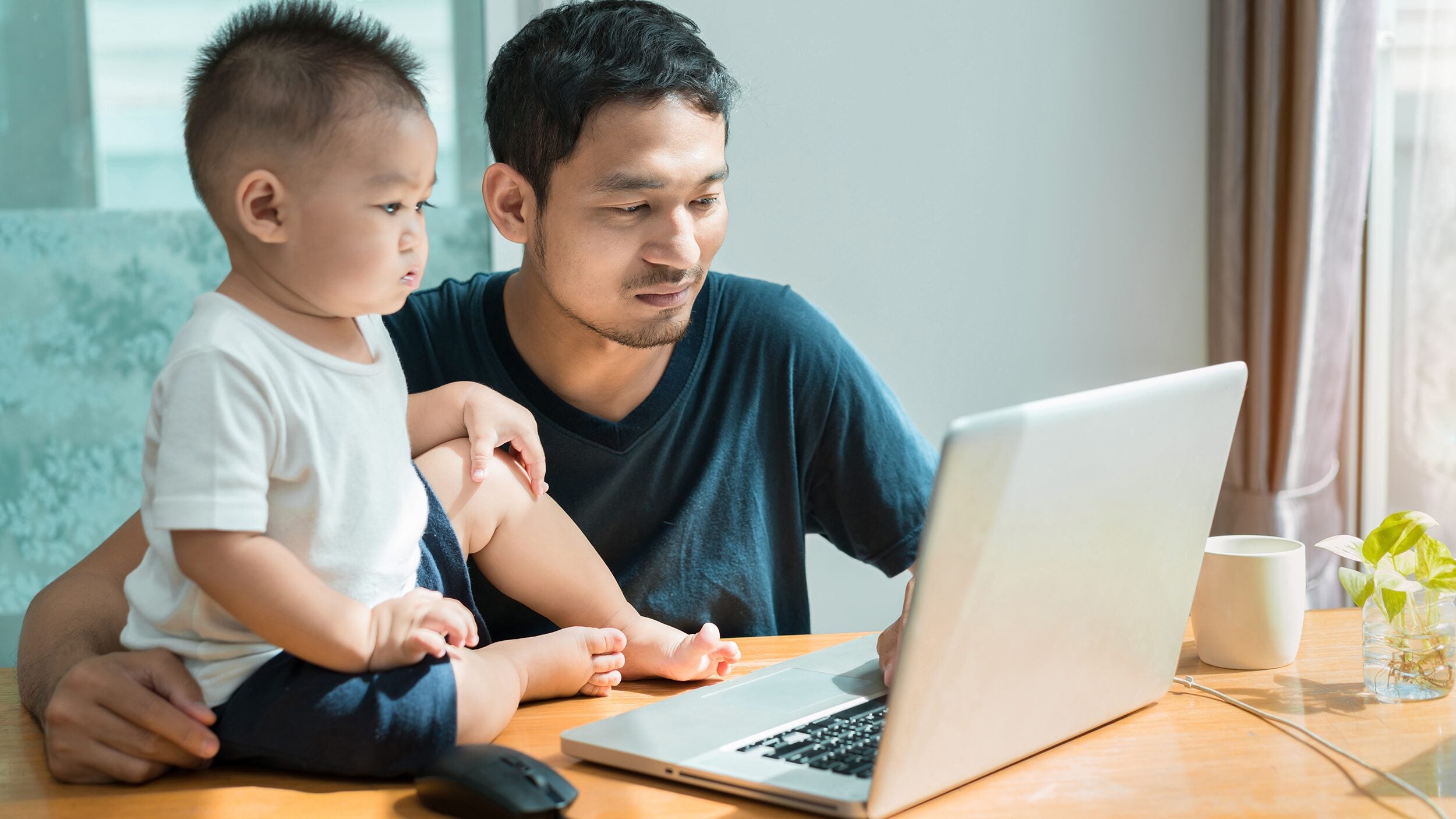 Man with a baby sitting at a table working on a laptop computer.